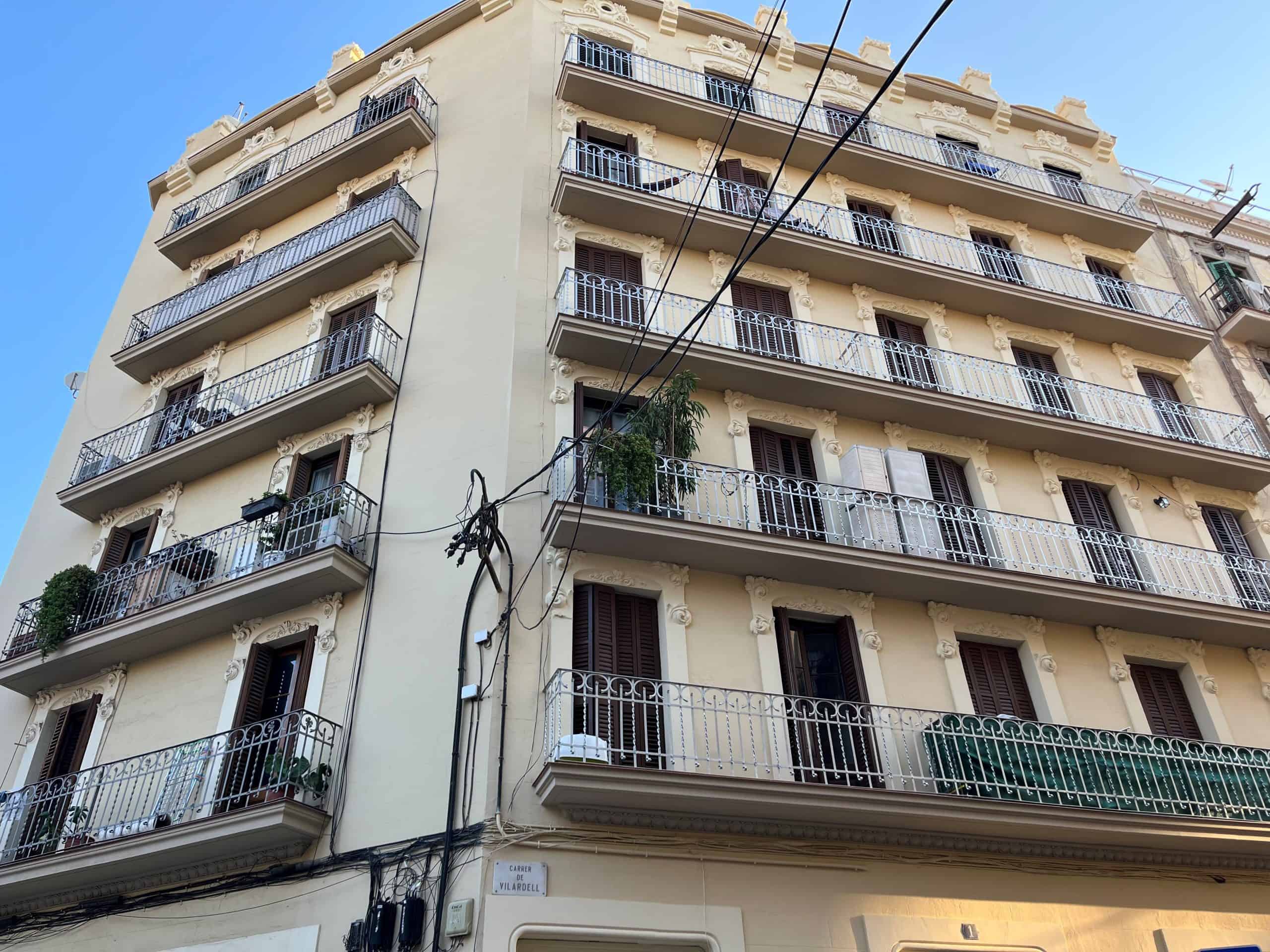 A multi-story beige building with ornate balconies and brown shutters, viewed from below against a clear blue sky. Electrical wires cross in front.