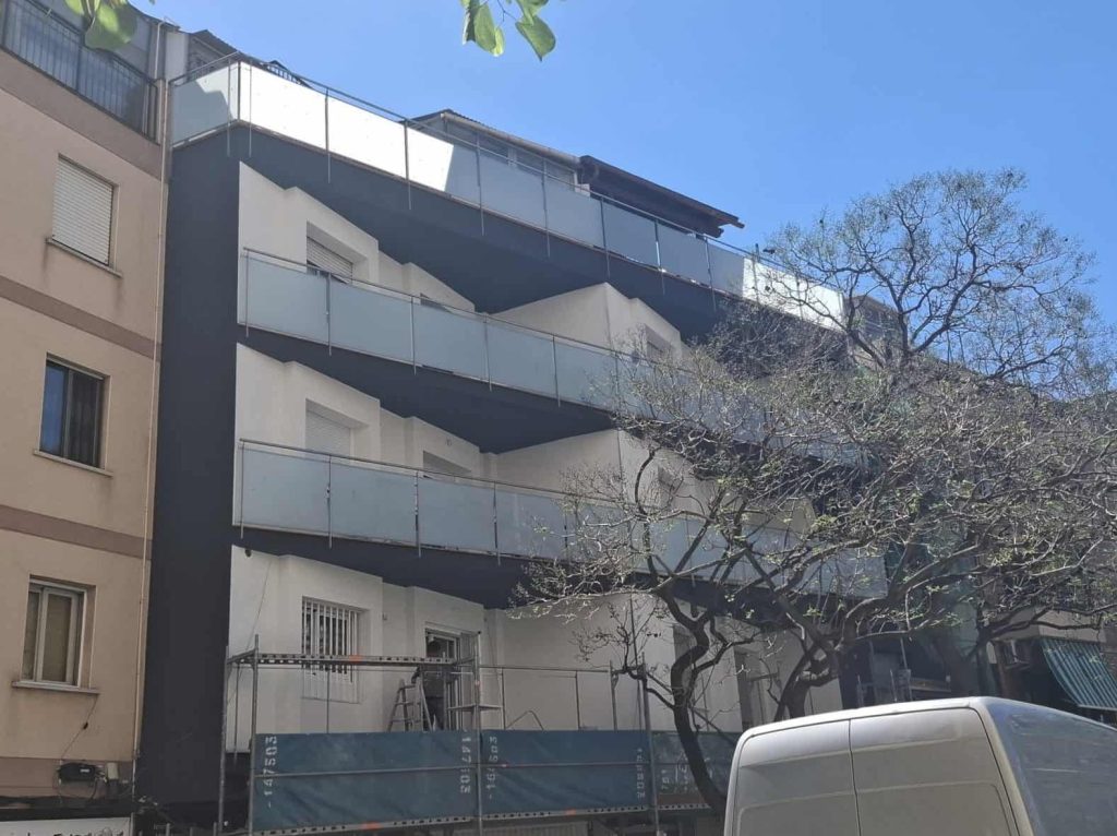 A four-story apartment building with scaffolding and construction workers on the balcony, next to another residential building, under a clear blue sky.