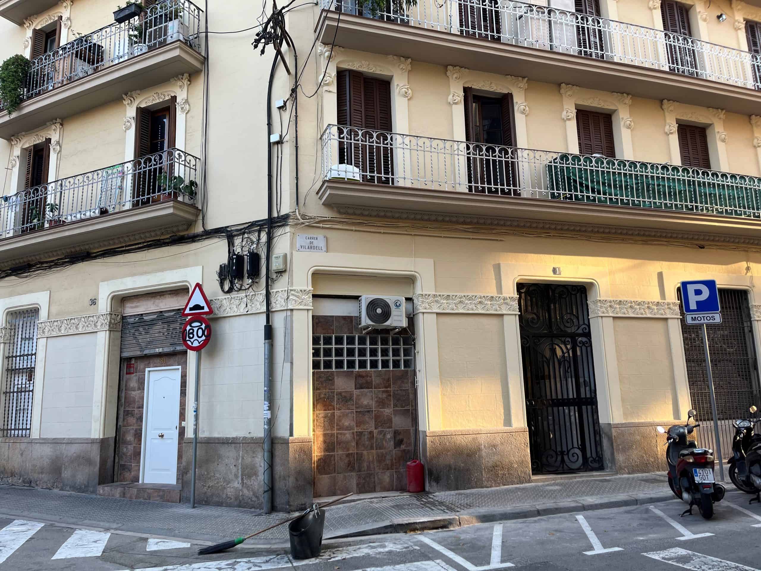 A corner building with balconies, closed shutters, a speed limit sign, a parking sign, and parked scooters on the street in front.