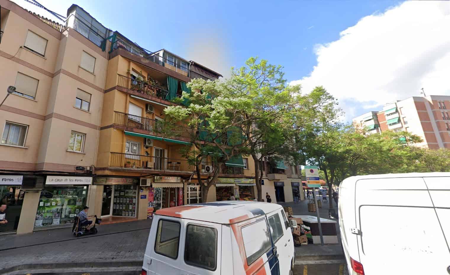 Street view of a multi-story apartment building with shops on the ground floor, parked vans, trees, and pedestrians on the sidewalk under a partly cloudy sky.