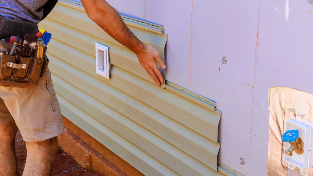 A person installs horizontal vinyl siding panels on the exterior wall of a house, with tools in a belt and plumbing fixtures visible.